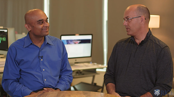 Two men are seated at a round table in an office setting, smiling and engaged in conversation. One man wears a blue shirt, and the other wears a dark shirt. Computer monitors are visible in the background.