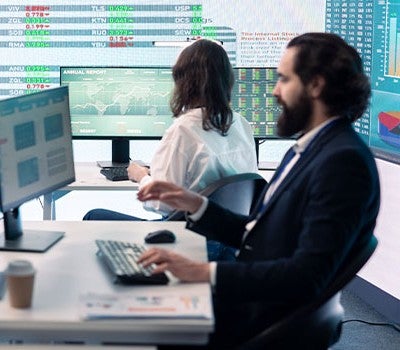 Two individuals seated at a desk, each working on their own computer, engaged in a collaborative environment.
