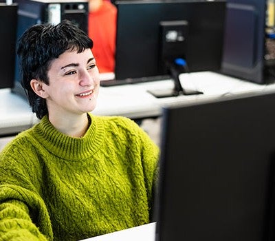 A woman in a green sweater smiles while seated at a computer, exuding a sense of joy and engagement.