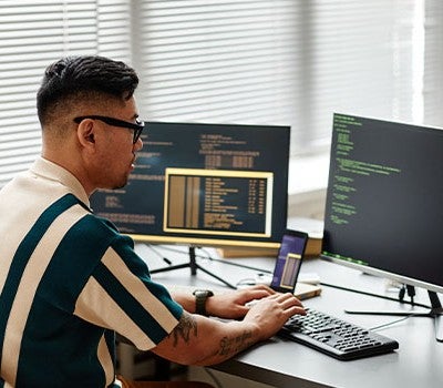 A man seated at a desk, focused on two computer screens displaying various tasks and information.