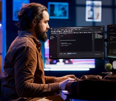 A man seated at a desk, focused on work, with two computer monitors displaying various tasks and information.