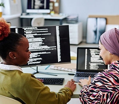 Two women at a desk with computers, focused on their work and collaborating on a project.