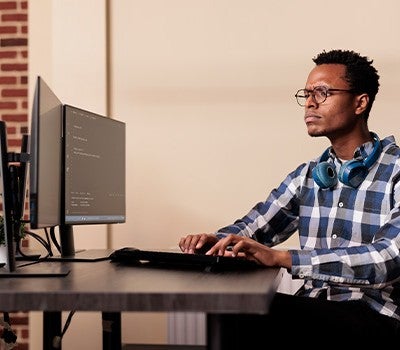 A man wearing headphones and a plaid shirt is seated at a desk, engaged with his computer.