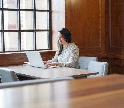 Woman working on a laptop at a table in a quiet study space with large windows and wood-paneled walls.